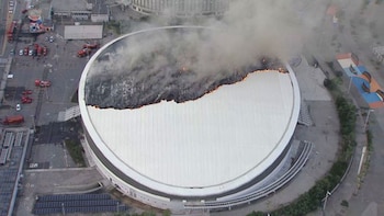 Vista aérea de un velódromo con parte de su techo blanco incendiado, emanando humo espeso, y varios vehículos de bomberos en el exterior