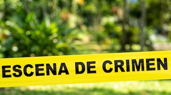 Cinta policial amarilla con letras negras que dicen "ESCENA DE CRIMEN" frente a un fondo desenfocado de árboles y vegetación verde.
