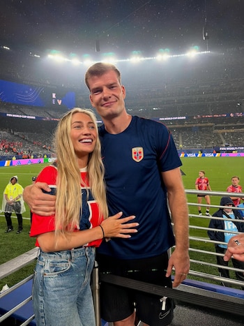 Un hombre alto y una mujer rubia sonriendo y abraz
dose en un estadio de f
tbol con luces y p
blico de fondo