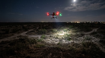 Dron en vuelo con luces rojas y verdes que ilumina el suelo en un terreno árido de noche con la luna de fondo.