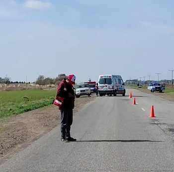 Vista lateral de una persona con gorra roja y chaqueta oscura en el arcén de una carretera. Al fondo, vehículos de emergencia y conos naranjas en el asfalto