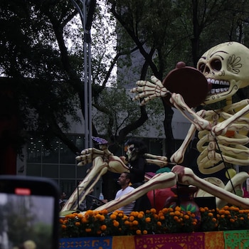Gran figura de esqueleto sobre un carro alegórico decorado con flores anaranjadas y personas a bordo en una calle con árboles