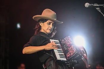 Una mujer con sombrero y camisa negra toca un acordeón negro con detalles rojos y teclas blancas en un escenario oscuro con luces brillantes