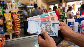 Manos sujetando billetes de la República Dominicana sobre una billetera negra frente a un mostrador con estantes de productos al fondo.