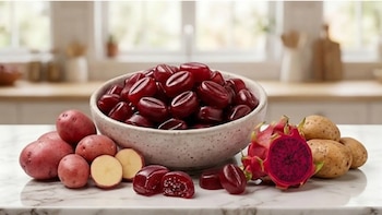 Un tazón lleno de gomitas rojizas en forma de grano de café sobre una encimera de mármol, flanqueado por papas rojas y amarillas, y una pitahaya abierta con pulpa morada