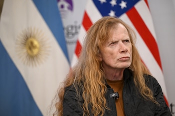 Primer plano de una persona con cabello largo rojizo-rubio, chaqueta negra y micrófono en la solapa, con banderas de Argentina y Estados Unidos difusas al fondo