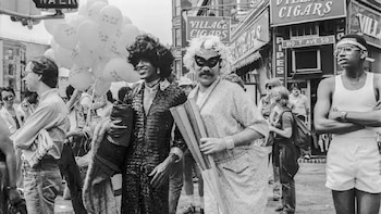 Una imagen en blanco y negro de Marsha P. Johnson sonriendo en un desfile, junto a otra persona enmascarada con peluca y un hombre joven al lado