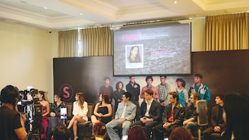 Vista de una conferencia de prensa con personas sentadas en primera fila, una pantalla proyecta información del equipo con foto de Gianella Neyra, y cámaras grabando el evento