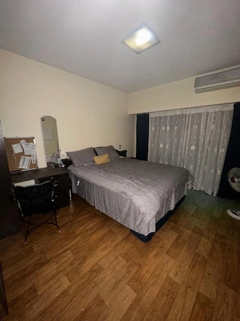 Dormitorio interior con cama doble gris, escritorio oscuro, silla negra, espejo y tablón de anuncios a la izquierda. Cortinas blancas y aire acondicionado visibles