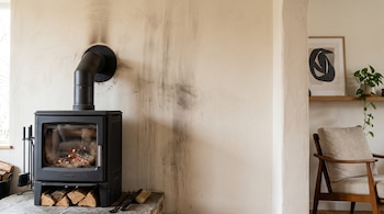 Estufa de leña negra encendida con leños debajo, marcas de hollín sobre la pared clara y un sillón visible a la derecha.