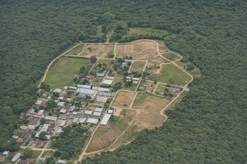 Vista aérea de un área boscosa que limita con un asentamiento de casas, edificios, caminos de tierra, campos verdes y parcelas de tierra seca.