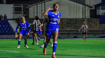 Una jugadora de fútbol con uniforme azul sonríe y se lleva la mano a la oreja en una cancha de césped. Hay otras jugadoras y asientos de estadio detrás.