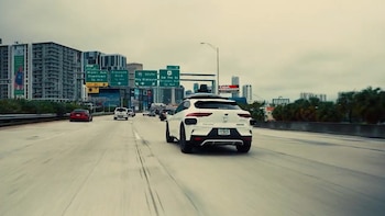 Vista trasera de un vehículo blanco de Waymo con equipo de sensores en el techo circulando por una autopista en Miami, con rascacielos al fondo y un cielo nublado