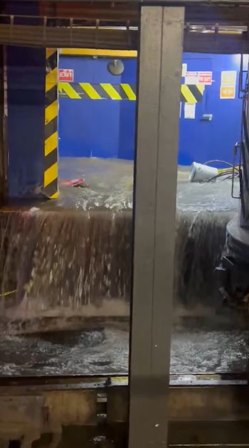 Agua fluye sobre un borde en un espacio interior con paredes azules, letrero de salida rojo, franjas de precaución amarillas y negras. Objetos flotan en el agua