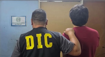 Vista de espalda de un agente con chaleco negro con la sigla DIC que sostiene el hombro de un hombre vestidocon camiseta roja