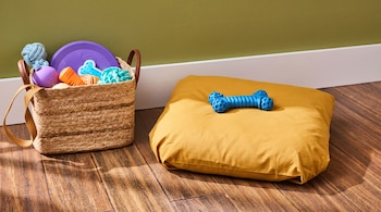 Una cesta con juguetes de colores para perros y una almohada amarilla con un hueso de plástico azul en el suelo de madera.