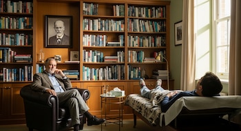 Un terapeuta barbudo sentado en un sillón de cuero frente a un paciente recostado en un diván, con una gran estantería de libros y un retrato de Freud detrás.