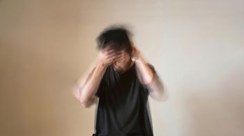 Una persona con camiseta oscura se cubre el rostro con las manos contra una pared clara, mostrando un marcado efecto de desenfoque de movimiento.