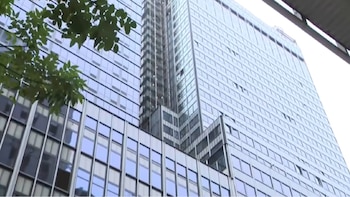 Un edificio moderno de muchos pisos con fachada de vidrio y ventanas cuadradas. Hojas verdes de un árbol aparecen en la esquina superior izquierda