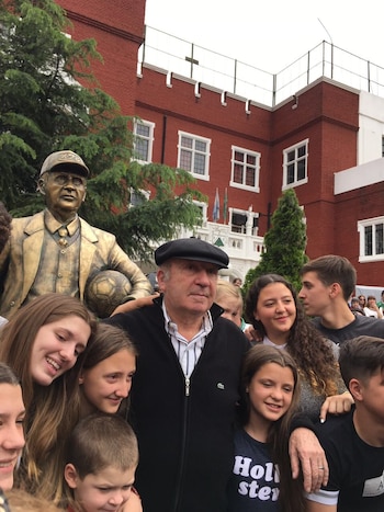 Un hombre mayor de boina negra junto a su estatua de bronce con balón, rodeado de varios jóvenes sonrientes, frente a un edificio de ladrillo rojo