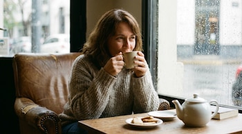 Una mujer con suéter gris toma de una taza en un sillón de cuero marrón junto a una ventana con gotas de lluvia, sobre una mesa hay una tetera y galletas.