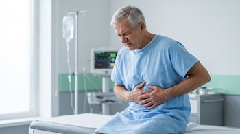Un hombre con bata azul sujeta su abdomen sobre una camilla médica frente a un soporte de suero y un monitor de signos vitales.