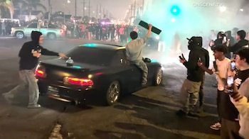 Una multitud observa un coche negro haciendo derrapes con humo y luces verdes en una calle oscura. Un hombre sentado en el techo levanta un objeto