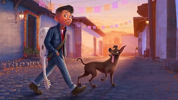 Niño con camisa roja, chaqueta de mezclilla y guitarra blanca camina junto a un perro por calle adoquinada con casas, papel picado y luz de atardecer