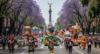 Vista aérea de un desfile en Paseo de la Reforma, CDMX, con carros alegóricos, personas en trajes tradicionales y jacarandás morados. El Ángel de la Independencia se alza al fondo.