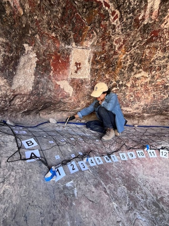 Mujer con gorra y guantes negros examina pared de cueva con pincel. Está sentada junto a una cuadrícula en el suelo. La roca del fondo exhibe arte rupestre