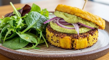 Primer plano de una hamburguesa dorada de plátano macho y quinoa con aguacate y cebolla morada, acompañada de una ensalada de hojas verdes en un plato de cerámica rústica.