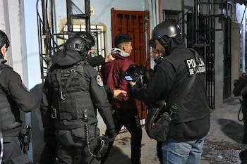 Varios agentes de policía con equipo táctico negro y cascos detienen a un joven con chaqueta roja y sudadera gris en una calle urbana
