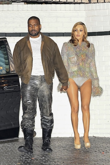 Kanye West, con chaqueta marrón, y Bianca Censori, con top multicolor y bragas, de la mano. Están sobre un suelo oscuro y una pared de ladrillo blanco