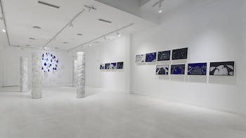Galería de arte blanca con columnas esculpidas, una instalación mural de gotas azules y obras abstractas enmarcadas en azul, blanco y negro