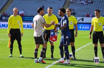 Dos jugadores de fútbol con uniformes diferentes se dan la mano en el campo verde, rodeados por árbitros con camisetas amarillas, previo a un partido