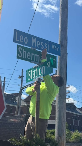 Un trabajador con camisa neón instala el letrero de la calle "Leo Messi Way" en un poste, junto a las señales de "Sherman St" y "Station"