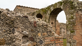 Primer plano de un muro de hormigón romano antiguo y erosionado con incrustaciones de piedras y ladrillos, y estructuras arqueadas borrosas al fondo.