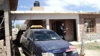 Automóvil azul con el parabrisas roto estacionado frente a una casa de ladrillo con un agente de uniforme junto a la puerta