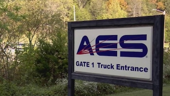 Primer plano de un letrero rectangular de AES GATE 1 Truck Entrance enmarcado en negro, rodeado de densa vegetación y árboles al fondo