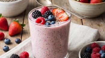 Vaso con bebida rosada y semillas de chía, cubierto con frambuesas, moras, arándanos y fresas, junto a una cuchara y tazones sobre madera.