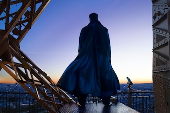 Hombre de espaldas con abrigo largo y gorra sobre una plataforma de vigas metálicas frente a una panorámica urbana al atardecer