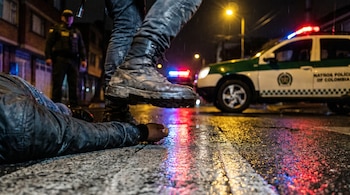 Primer plano de una bota de combate pisando cerca de una persona en el suelo, con un coche patrulla de la Policía Nacional de Colombia y un agente borrosos al fondo.