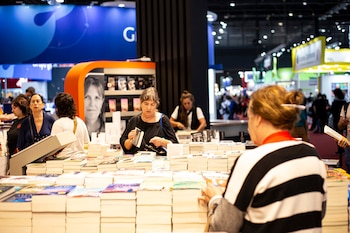 Varias personas, mayormente mujeres, navegan entre grandes pilas de libros en mesas y estanterías dentro de un recinto de exposición bien iluminado
