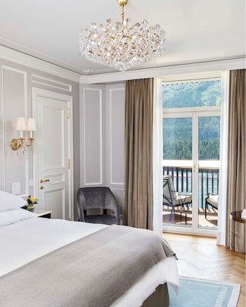 Cama, manta, mesa de noche con flores, silla, candelabro de cristal, aplique de pared, puerta, ventanal y balcón. Montañas y lago al exterior