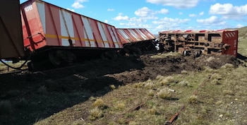 Tres vagones de carga rojos con franjas blancas volcados sobre la tierra a un costado de las vías del tren