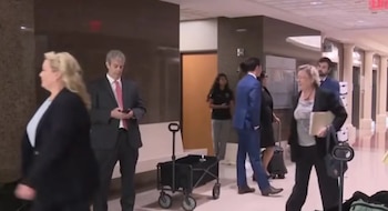 Personas caminan por un pasillo interior, algunas vestidas de traje, una mujer rubia de perfil, un hombre mirando su teléfono, y un carrito plegable negro