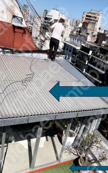 Un hombre de espaldas de pie sobre un tejado de chapa acanalada con cables. Alrededor se ven edificios, y abajo, un balcón con plantas en macetas