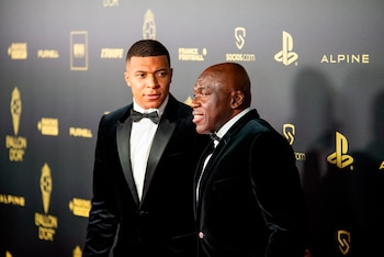 Kylian Mbappé y Wilfried Mbappé, ambos vestidos con esmoquin, posan juntos frente a un fondo oscuro con logotipos dorados de patrocinadores de fútbol