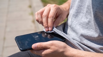 Manos de una persona que sostienen un teléfono móvil y limpian la pantalla de vidrio con la tela de una camiseta gris.