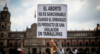Mano sostiene cartel blanco con texto negro sobre aborto por violación en Tamaulipas. Fondo con edificio histórico y bandera de México borrosos.